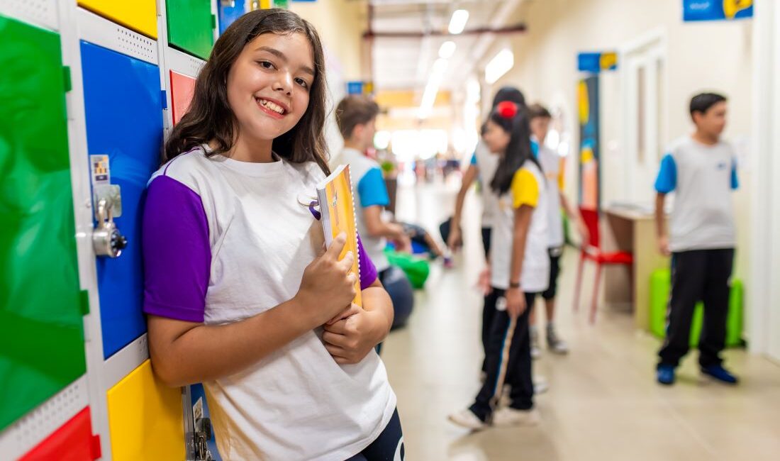 Estudante do sexo feminino recém-saída do período de alfabetização encostada em um armário no corredor.