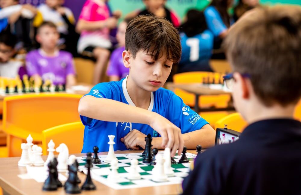 Menino estudante de camisa azul jogando xadrez em um ambiente com outros alunos jogando.