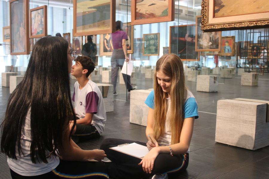 Na imagem, três jovens estão dentro de um museu ou galeria de arte, que exibe várias obras penduradas em paredes de vidro ao fundo. À esquerda, uma garota com longos cabelos negros, lisos e soltos está sentada na direção diagnonal para um garoto, que usa uma camiseta branca com detalhes roxos e olha para cima, possivelmente em direção a uma das obras de arte ou conversando com a garota. À direita, uma garota loira, sentada e olhando para baixo, está escrevendo ou desenhando em um caderno sobre uma prancheta. Ela usa uma camiseta azul e tem um cabelo longo e liso. O ambiente tem um piso escuro e é iluminado por luz natural, sugerindo um espaço amplo e moderno, propício para a aprendizagem baseada em projetos.