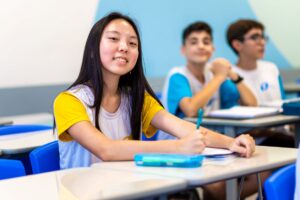 Estudante asiática sentada em carteira escolar olhando e sorrindo em direção à câmera.