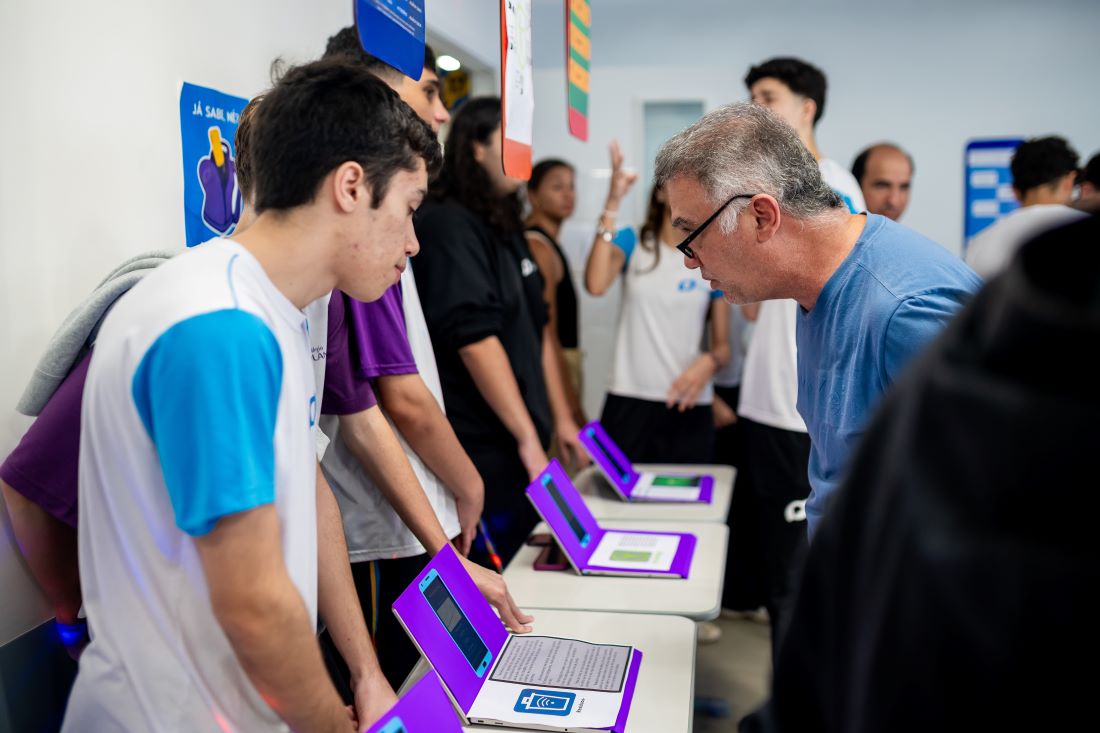 A imagem retrata um ambiente educacional, possivelmente uma feira ou exposição de projetos. No centro, um homem mais velho, de óculos e camiseta azul, observa atentamente um estudante. O jovem veste uma camiseta branca com mangas azuis e fica ao lado de outros colegas, interagindo com materiais expostos em mesas. As mesas contêm pastilhas com molduras roxas e documentos descritivos, indicando a apresentação de trabalhos ou ideias. Ao fundo, outros estudantes e adultos participam do evento. Este cenário ilustra a prática da Educação empreendedora , promovendo o protagonismo estudantil e o desenvolvimento de habilidades inovadoras e colaborativas.
