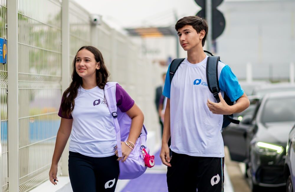 Dois estudantes com uniforme escolar caminham lado a lado na calçada próxima à entrada da escola carregando mochilas.