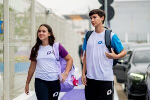 Dois estudantes com uniforme escolar caminham lado a lado na calçada próxima à entrada da escola carregando mochilas.