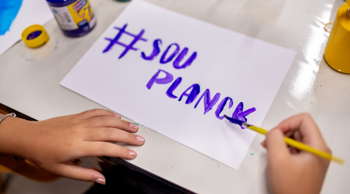 Estudante pinta "#SOU PLANCK" com tinta roxa em atividade criativa que integra a metodologia de ensino do Colégio Planck.