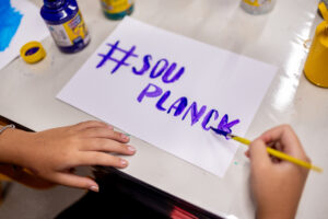 Estudante pinta "#SOU PLANCK" com tinta roxa em atividade criativa que integra a metodologia de ensino do Colégio Planck.