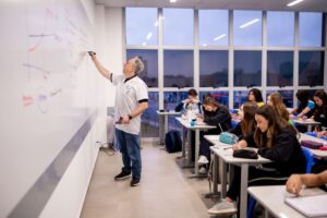Professor do Colégio Planck explica conceitos de matemática no quadro branco durante aula interativa com estudantes do Ensino Fundamental II e Médio.