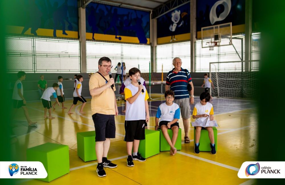Pai e filho em apresentação no Dia da Família Planck na quadra poliesportiva, com avô e outros estudantes participando do evento que fortalece vínculos entre família e colégio.