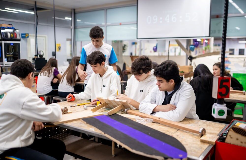 Estudantes realizam atividade prática em grupo em laboratório escolar, utilizando ferramentas e materiais de madeira sobre uma mesa.