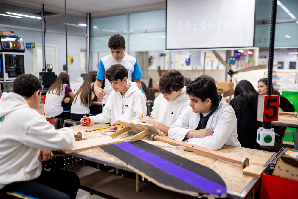 Estudantes realizam atividade prática em grupo em laboratório escolar, utilizando ferramentas e materiais de madeira sobre uma mesa.