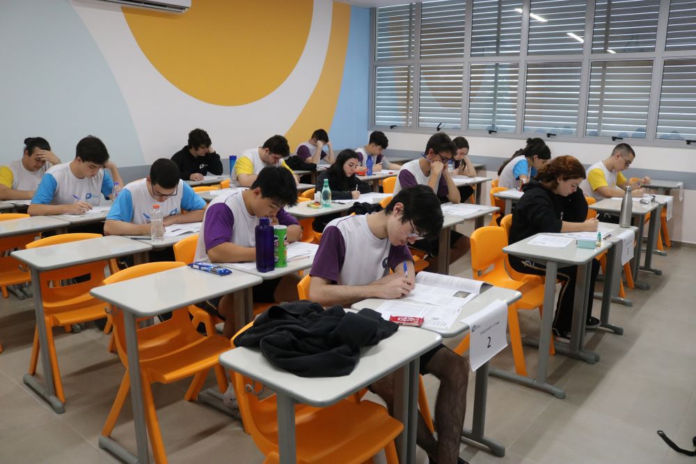 Estudantes do Colégio Planck concentrados em avaliação estruturada, escrevendo em carteiras individuais durante simulado, demonstrando disciplina, foco e constância no ambiente de aprendizado organizado.
