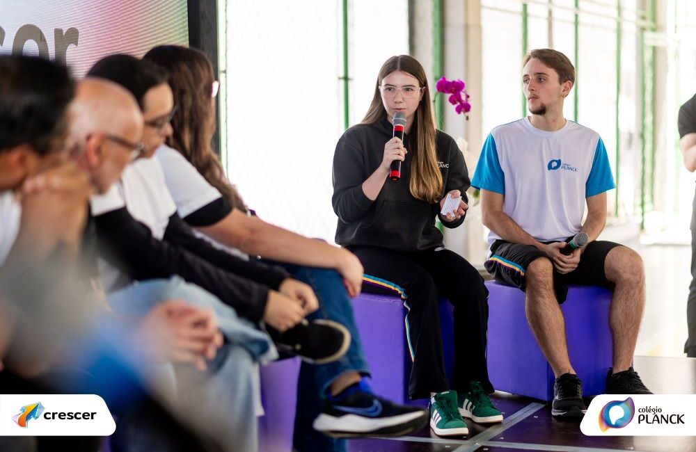 Autonomia se aprende: jovem fala ao microfone em roda de conversa escolar, acompanhada por colega e público atento.