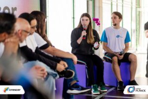 Autonomia se aprende: jovem fala ao microfone em roda de conversa escolar, acompanhada por colega e público atento.