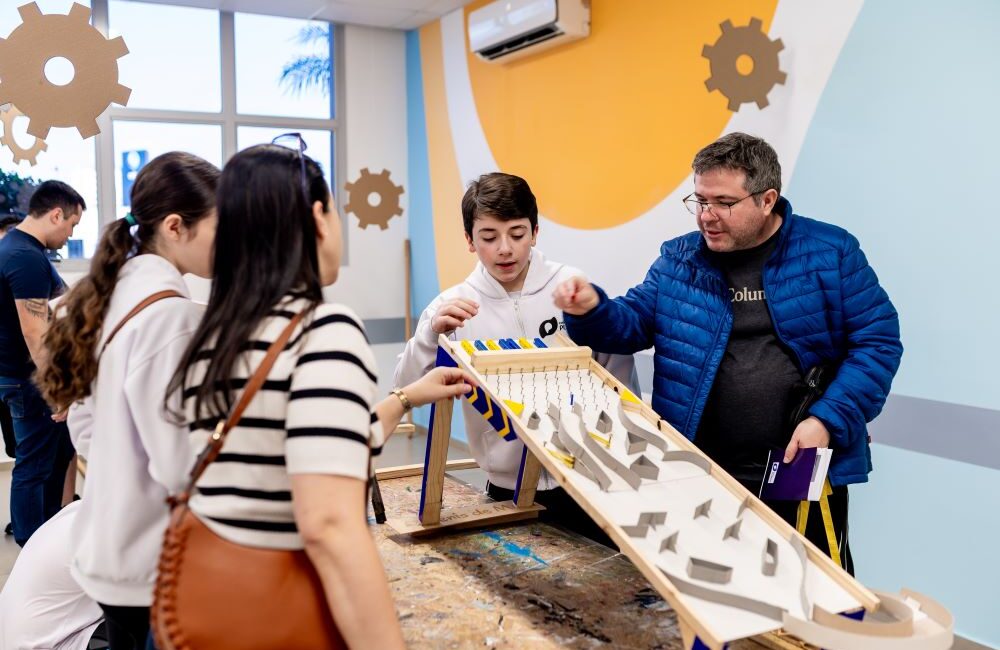 Estudante apresenta maquete interativa para grupo em ambiente escolar, com adultos acompanhando a explicação.