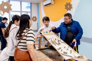 Estudante apresenta maquete interativa para grupo em ambiente escolar, com adultos acompanhando a explicação.