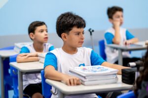 Estudantes uniformizados atentos à aula, sentados em carteiras com livros sobre a mesa.