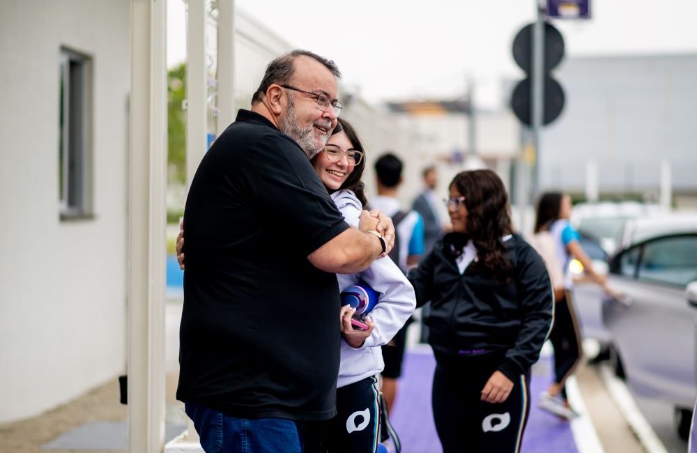 Professor abraça aluna adolescente na entrada da escola enquanto outros estudantes passam pela calçada ao fundo, em momento de despedida antes das aulas.