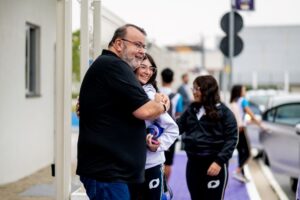 Professor abraça aluna adolescente na entrada da escola enquanto outros estudantes passam pela calçada ao fundo, em momento de despedida antes das aulas.