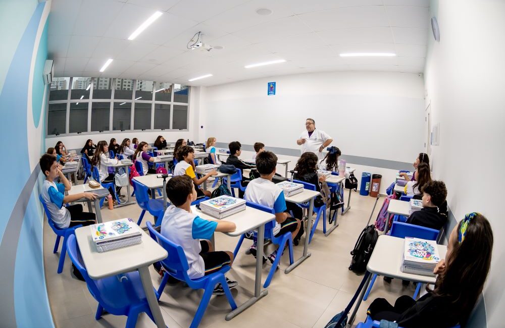 Professor explica conteúdo para estudantes sentados em sala de aula organizada, com estudantes usando uniforme e acompanhando a aula.