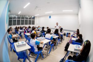 Professor explica conteúdo para estudantes sentados em sala de aula organizada, com estudantes usando uniforme e acompanhando a aula.