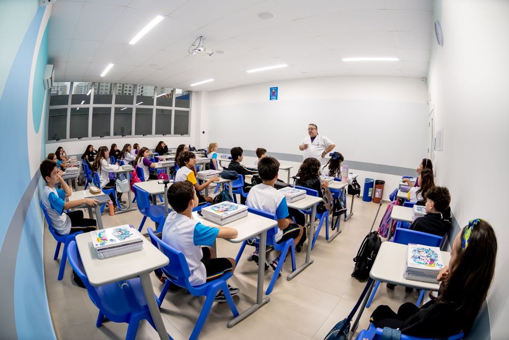 Professor explica conteúdo para estudantes sentados em sala de aula organizada, com estudantes usando uniforme e acompanhando a aula.

