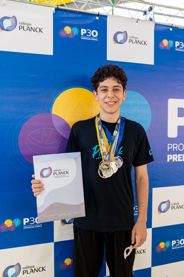 Marcos Birolini aparece sorrindo em evento do Colégio Planck, segurando certificado e usando medalhas, representando sua conquista de bolsa integral na Universidade de Toronto.