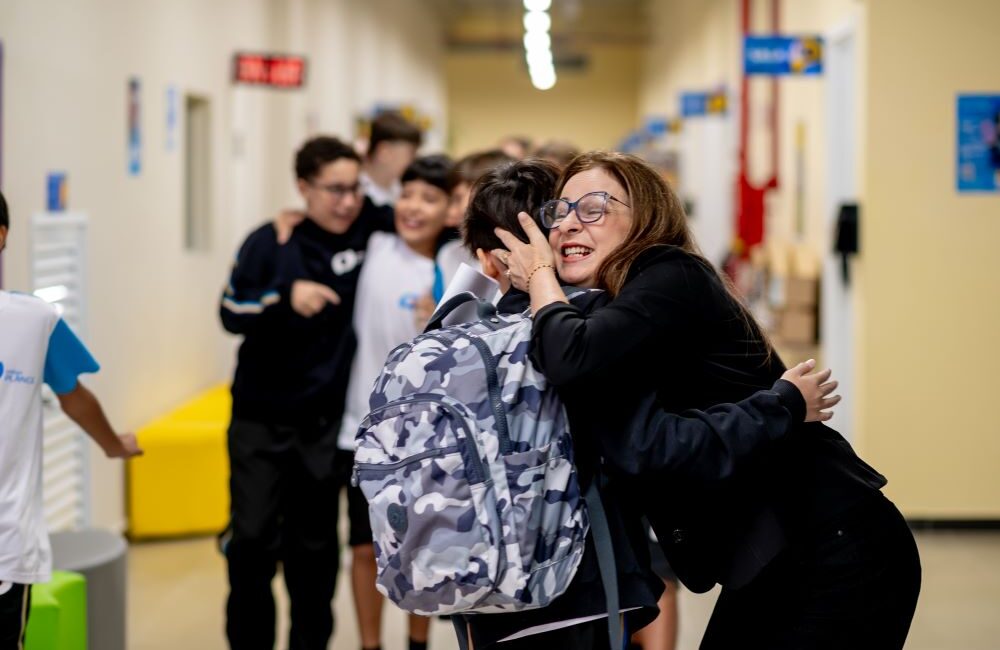 Educadora abraça estudantes em corredor escolar, representando como o 6º ano é uma mudança acadêmica ou emocional na adaptação dos estudantes.