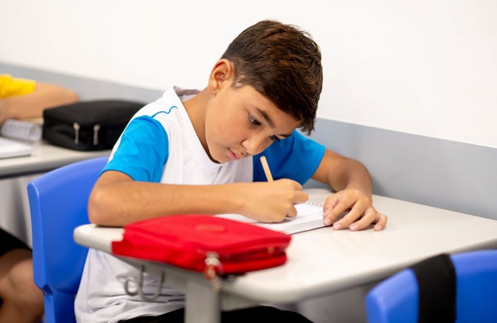 Estudante concentrado escreve em um caderno durante a aula, representando um processo de aprendizagem baseado em desafios, raciocínio e desenvolvimento do pensamento crítico.