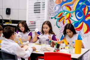 Crianças participam de uma oficina criativa utilizando materiais diversos para desenvolver projetos, representando a criatividade como uma habilidade treinável e essencial para o futuro.