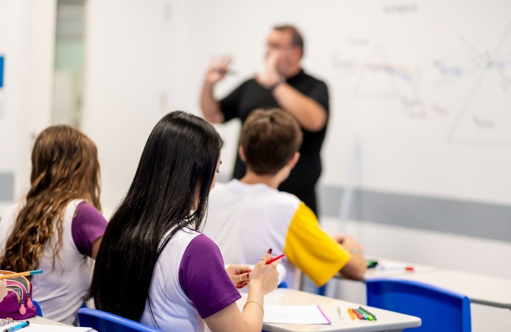 Estudantes acompanham a explicação de um professor em sala de aula, com cadernos e materiais escolares sobre as carteiras, representando o desenvolvimento de habilidades no Ensino Médio para além do vestibular.