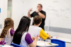 Estudantes acompanham a explicação de um professor em sala de aula, com cadernos e materiais escolares sobre as carteiras, representando o desenvolvimento de habilidades no Ensino Médio para além do vestibular.