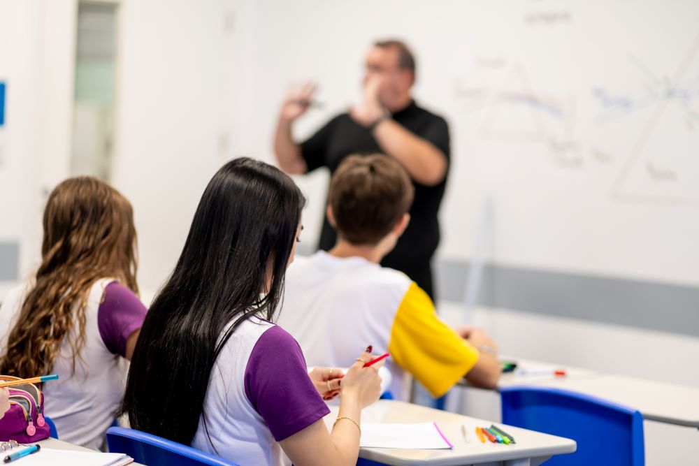 Estudantes acompanham a explicação de um professor em sala de aula, com cadernos e materiais escolares sobre as carteiras, representando o desenvolvimento de habilidades no Ensino Médio para além do vestibular.